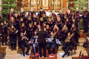 Abb. der Chorgemeinschaft Emmersdorf vor dem Hochaltar der Pfarrkirche St. Nikolaus in Emmersdorf an der Donau. Vor dem Chor ein Bläserensemble, im Vordergrund mit dem Rücken zum Publikum der Dirigent Florian Neilinger.